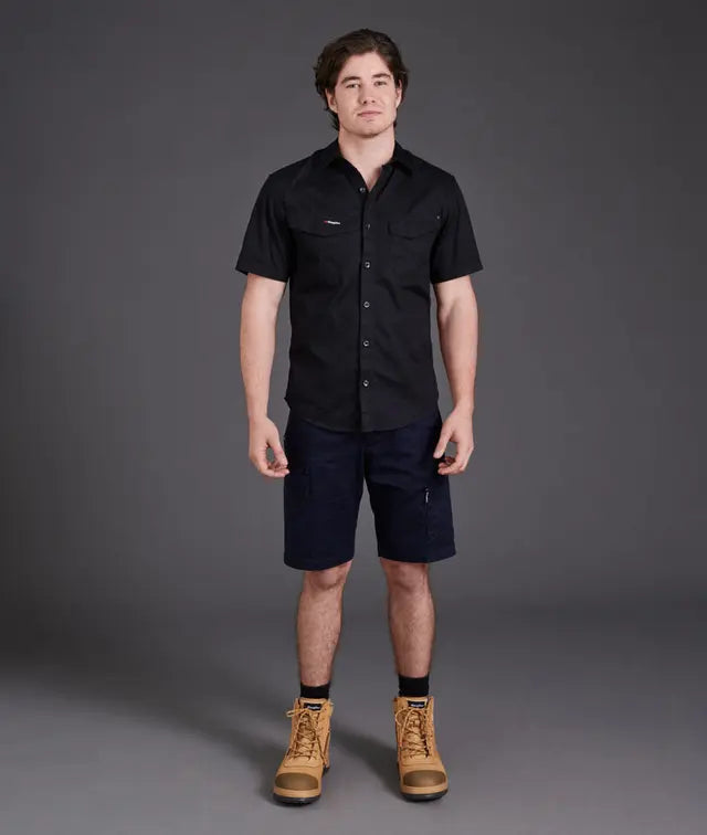 Man wearing a black shirt, navy shorts, and brown boots against a gray background
